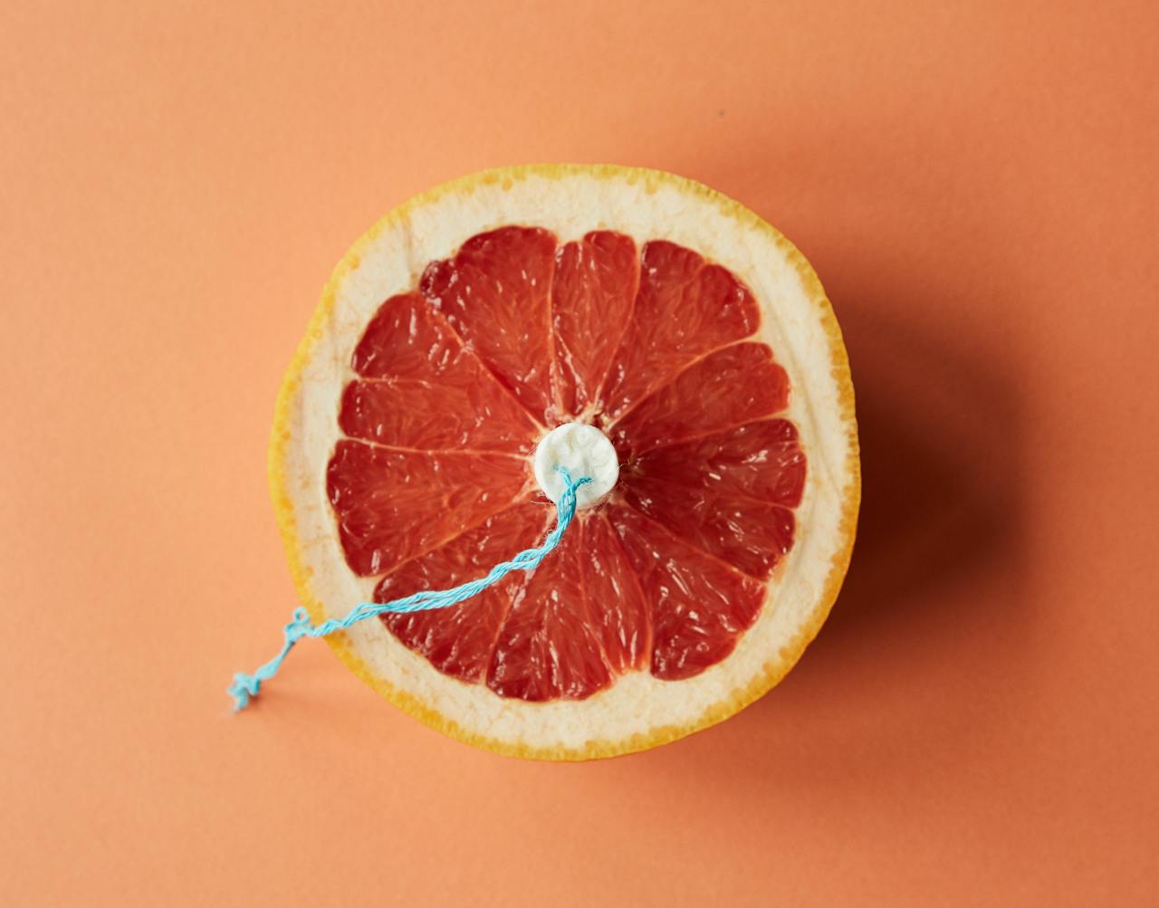 Artistic depiction of menstrual theme with grapefruit and tampon on orange background.